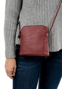 Bolso de hombro de cuero burdeos con cierre de cremallera, forma rectangular, textura suave y detalle de logo grabado en la parte inferior.