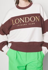 Persona vestida con un sudadera blanca y marrón con el texto "LONDON Leisure Club" y pantalones verdes brillantes, con las manos en los bolsillos, contra un fondo liso.