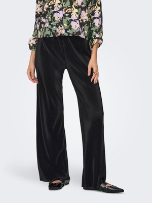 Torse et jambes d'une personne portant un pantalon noir plissé à jambes larges, un chemisier fleuri à manches longues, des ballerines noires pointues et une bague en or à la main droite.