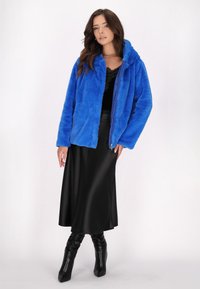 Giacca in pelliccia sintetica blu brillante con cappuccio, dotata di chiusura con zip. Abbinata a una gonna nera di raso e stivali neri al ginocchio.