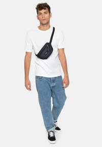 Svart canvas axelväska med dragkedja, bärs diagonalt över en vit t-shirt och blå jeans, och matchas med svartvita sneakers.