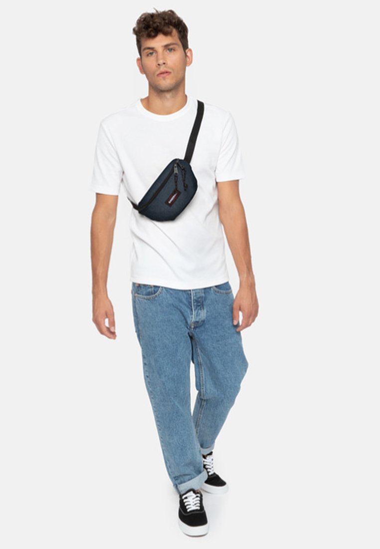 Svart canvas axelväska med dragkedja, bärs diagonalt över en vit t-shirt och blå jeans, och matchas med svartvita sneakers.