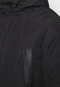 Svart puffjacka med huva, dragkedjestängning och textilstruktur med en slät accentpanel på sidan.