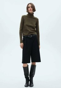 Olijfgroene coltrui, zwarte high-waisted wijde shorts en zwarte kniehoge laarzen. Gladde textuur en getailleerd ontwerp.