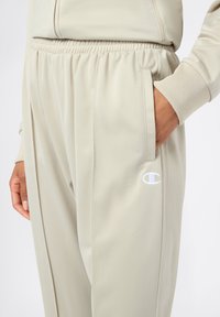 Pantaloni sportivi beige chiaro con vita elastica e tasche laterali. Presentano un logo ricamato bianco sulla coscia sinistra. Texture liscia.