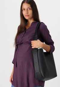 Borsa tote in pelle nera con forma strutturata, ampia tracolla e texture liscia. Il modello indossa una tunica viola annodata in vita.