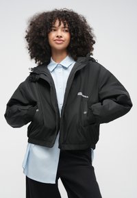 Čierna krátka bunda so zapínaním na zips, elastickými manžetami a nastaviteľnou kapucňou, nosená cez svetlomodrú košeľu s priliehavým golierom. Hladká textúra.