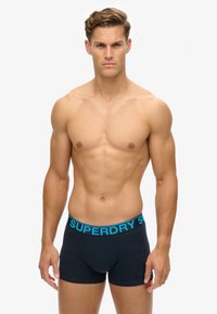 Czarne bokserki z niebieskim paskiem "SUPERDRY", charakteryzujące się dopasowanym krojem i gładką teksturą materiału. Model stoi z rękami swobodnie opuszczonymi wzdłuż ciała.