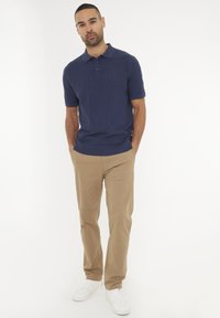 Námořnické polo tričko s krátkým rukávem a texturovaným vzorem, spárované s béžovými kalhotami a bílými teniskami, stojící na jednobarevném pozadí.