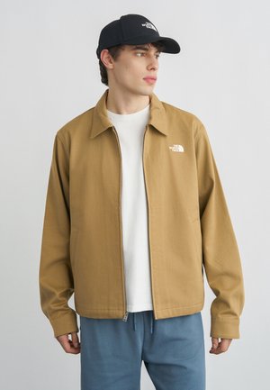 Jonge man die een beige jack met ritssluiting en een zwarte pet met The North Face-logo's draagt, een wit shirt en een blauwe broek, en naar rechts kijkt.