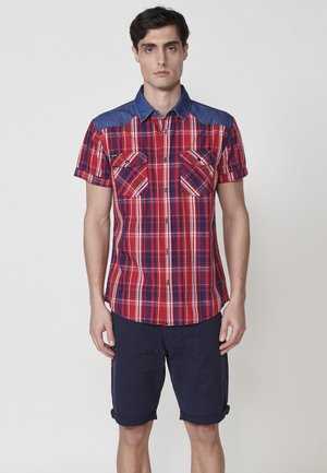 Cămașă cu mâneci scurte în carouri roșii și bleumarin, cu guler din denim, având două buzunare pe piept și un tiv curbat. Pur și simplu cu pantaloni scurți bleumarin răsuciți.