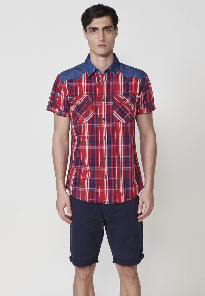 Kurzarmhemd in rot-blauem Karomuster mit Denimkragen, ausgestattet mit zwei Brusttaschen und einem abgerundeten Saum. Kombiniert mit navyfarbenen, hochgekrempelten Shorts.