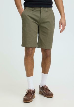 Mann iført olivengrønne shorts, hvite sokker og brune skinnbåtsko, stående med den ene hånden i lomma mot en ensfarget bakgrunn.