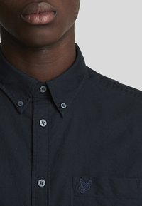 Camisa de un oscuro color azul marino con cuello abotonado, que presenta un pequeño logo bordado en el bolsillo del pecho y una tela con textura sutil.