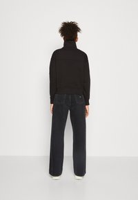 Felpa nera con colletto alto, abbinata a jeans in denim scuro a gamba larga. Il tessuto sembra liscio; dettagli delle cuciture visibili sulle spalle.