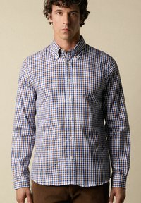 Homme portant une chemise à carreaux bleue, brune et blanche à manches longues sur un fond beige uni.