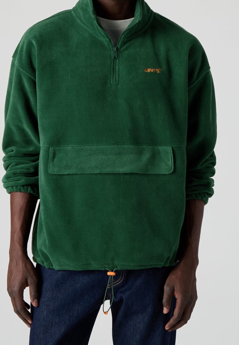 Personne portant un pull polaire vert avec un demi-zip, une grande poche sur le devant et le logo Levi’s orange, associé à un jean foncé.