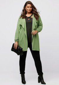 Hellgrüner, offener Cardigan mit langen, elastischen Ärmeln, kombiniert mit einem schwarzen Top und schmalen schwarzen Hosen. Schwarze Clutch und Stiefel mit Absatz.