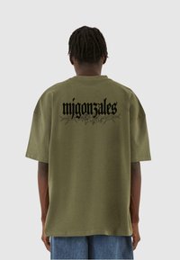 Oliven grøn oversized t-shirt i bomuld, med sort tekst "migonzales" og blomsterdesign på ryggen. Korte ærmer, afslappet pasform.
