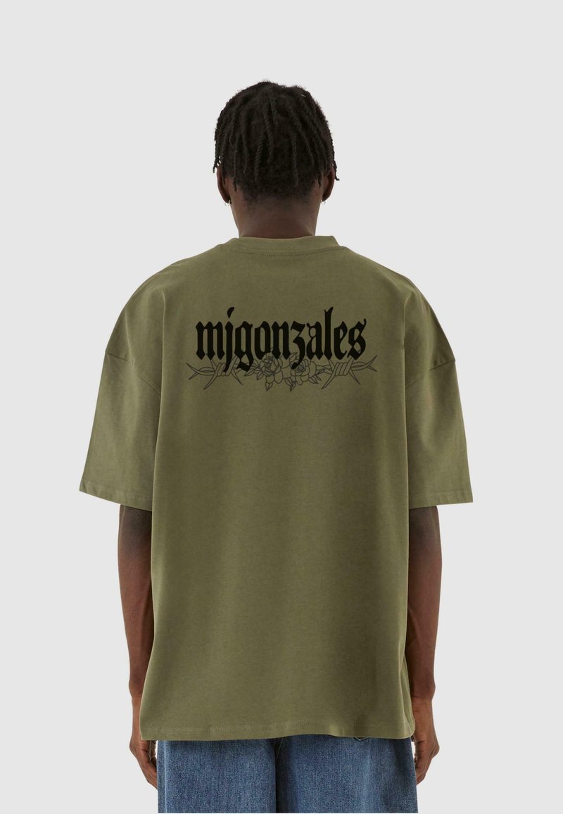 Oliven grøn oversized t-shirt i bomuld, med sort tekst "migonzales" og blomsterdesign på ryggen. Korte ærmer, afslappet pasform.