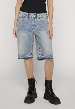 Persoană purtând pantaloni scurți din denim bleu deschis până la genunchi, cu tivuri destrămate, cizme negre cu platformă și șiret și un top negru.