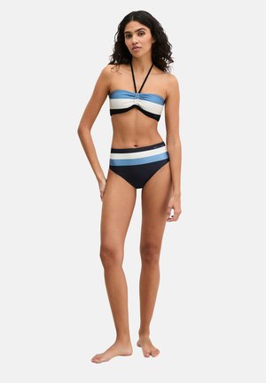 Frau steht barfuß und trägt einen zweiteiligen Badeanzug mit blauen, weißen und schwarzen Streifen und Neckholder-Oberteil vor einem schlichten weißen Hintergrund.