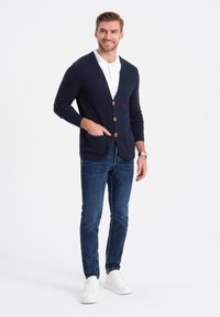 Cárdigan navy con cuello en V, botones de madera y dos bolsillos delanteros, llevado sobre una camiseta polo blanca, combinado con jeans azules y zapatillas blancas.