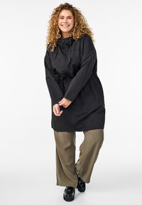 Schwarze Zip-Jacke mit verstellbarem Kordelzug an der Kapuze und verstellbarem Bund, ausgestattet mit vorderen Taschen und einem lockeren Schnitt, kombiniert mit olivgrünen Hosen.