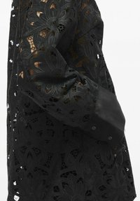 Robe en dentelle noire ornée de motifs floraux avec des détails ajourés. Manches longues avec un poignet noir uni et une fermeture à bouton.