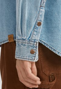 Light blue denim shirt with long sleeves, buttoned cuffs, and brown leather tag. Worn with textured brown corduroy pants.