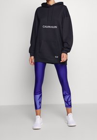 Felpa nera oversize con logo "CALVIN KLEIN", tasca frontale, abbinata a leggings viola con design lucido e pattern, e sneakers bianche.
