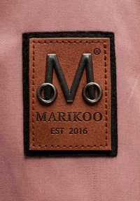 Hnedá kožená nášivka s vyrazeným čiernym logom „M“, obklopená čiernym okrajom z filcu, s textom „MARIKOO EST 2016“ pod ním na svetloružovej látke.