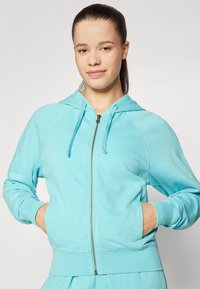 Svijetloplava zip-up dukserica s rebrastim završetkom i manžetama, sa kapuljačom i džepovima s obje strane, izrađena od mekog, teksturiranog materijala.