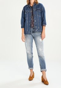 Giacca di jeans con vestibilità comoda, colore blu scuro, tasche, indossata sopra un top con fantasia. Jeans chiari strappati e mule con tacco marroni.