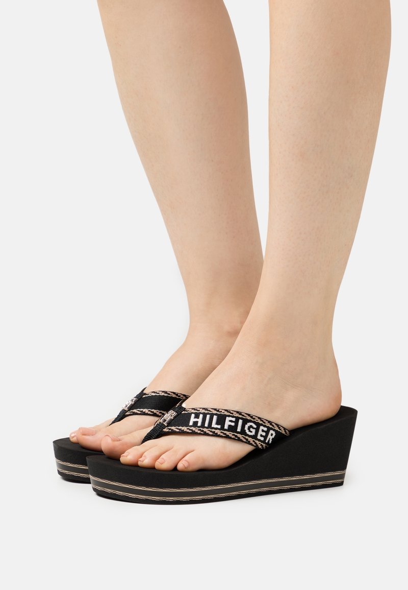 Pieds portant des tongs compensées noires avec "HILFIGER" sur les brides, sur un fond clair uni.