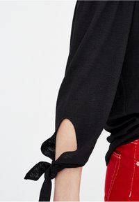 Schwarze Bluse mit Dreiviertelärmeln, die Ausschnittdetails und gebundene Akzente an den Manschetten aufweist, aus einem leichten, strukturierten Stoff gefertigt.