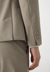 Primo piano della mano di una persona vicina al fianco, che indossa un completo beige strutturato con tre bottoni sul manica della giacca.