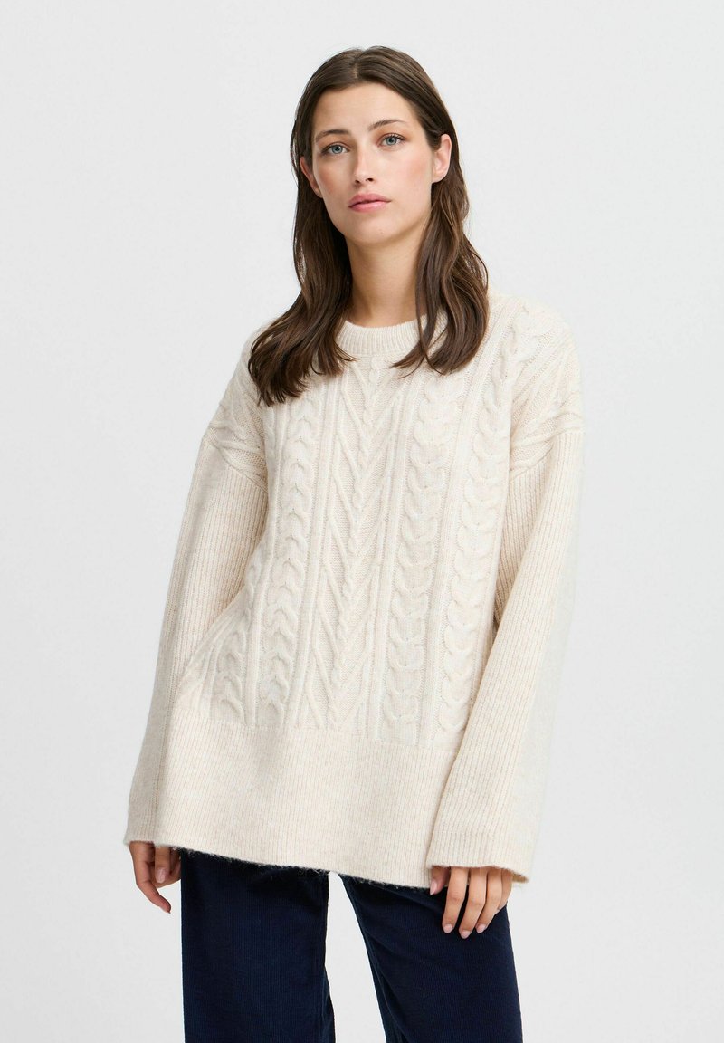 Pull en laine crème avec motif en maille torsadée, offrant une coupe décontractée, des poignets larges en côtes et un motif texturé sur l'ensemble. Matière douce et pelucheuse.
