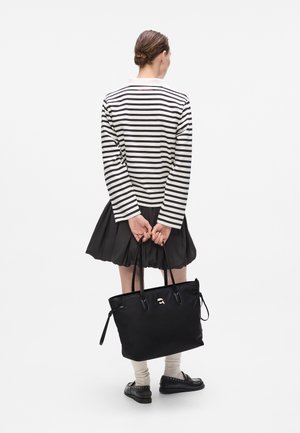 Borsa tote nera con texture liscia, dotata di due manici e un accento logo argentato. Indossata una camicia a righe con maniche lunghe e una gonna nera plissettata.