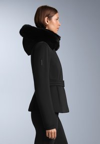 Crna jakna s kapuljačom obloženom krznom, krojenog fit-a, detaljem remena i strukturiranim dizajnom. Ima glatku teksturu i elegantnu siluetu.