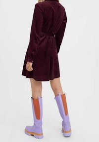 Vestito in velluto a coste di un profondo bordeaux con vita cinturata e maniche lunghe, abbinato a stivali alti fino al ginocchio di colore lavanda con dettagli arancioni.