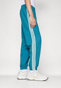 Persona in pantaloni jogging color teal con strisce triple bianche e sneakers bianche che si trova accanto a uno sfondo semplice.