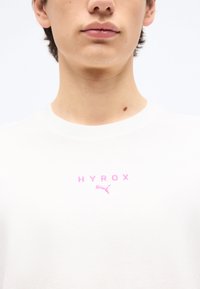 Νεαρό άτομο που φορά λευκό πουκάμισο με ροζ κείμενο «HYROX» και λογότυπο Puma στο κέντρο του στήθους, με απλό φόντο.