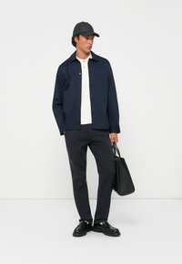 Navy button-up jacket over a white polo, dark pants, black shoes, and a black cap, holding a black bag. Smooth fabric and structured design.