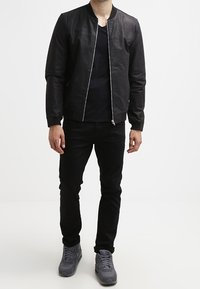 Mand iført sort bomberjakke, sorte bukser, sort t-shirt og grå sneakers, som står mod en hvid baggrund.