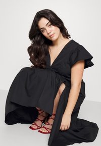 Black, knee-length dress with a deep V-neck and ruffled sleeves. Features a flowy skirt with a side slit. Paired with red strappy heels.