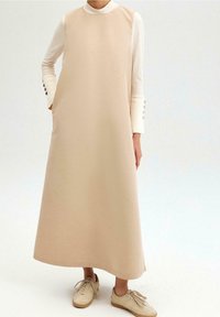 Beige mekko ilman hihoja, jossa on korkea kaula-aukko, sileä pinta ja suora siluetti. Päällä pitkähihaisen valkoisen paidan, jossa on nappikoristeet.