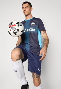 Camisa de futebol azul e azul-marinho com logotipo e texto, combinada com calções azul-marinho e meias brancas. O jogador equilibra uma bola de futebol no joelho.