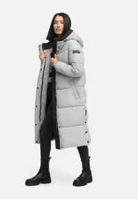 Grå, knælang pufferjakke med hætte, der har en lynlås, trykknapper og elastiske manchetter. Båret over sorte tøj og støvler.