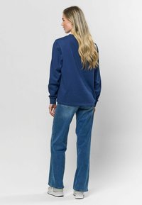 Jūras zila džemperis ar garām piedurknēm, brīvs griezums un ribotas aproces, apvienots ar augstās jostasvietas ziliem džinsiem un baltiem sporta apaviem.